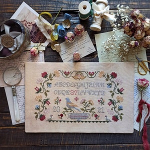 Puede incluir: Un sampler de punto de cruz con un fondo beige claro que presenta un pájaro azul posado en una rama con flores rosas, rojas y amarillas. El alfabeto está bordado en rosa y los números 20 y 21 están bordados en rojo.