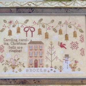 May include: Framed cross-stitch artwork with a Christmas theme. The design features a house, snowman, bells, and the text "Caroling, caroling, Christmas bells are ringing!" in a rustic style. The frame is a light color with a dark brown inner border.