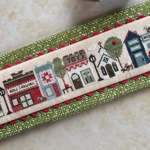 May include: Cross-stitch sampler depicting a quaint village scene with shops and buildings. The sampler features buildings in red, blue, and brown, with the text "Kiki Shopping" visible. The sampler is framed with a red scalloped edge and rests on a green patterned fabric.