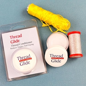 May include: An assortment of sewing supplies on a teal surface. Includes a package of "Thread Glide" thread conditioner, two containers of thread glide, a spool of silver thread with red ends, and a yellow thread. The text on the package reads "Reduces Tangles And Fraying."