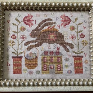 May include: Cross-stitch artwork in a cream-colored frame. The design features a brown rabbit leaping over a house and basket of eggs. Floral and alphabet details surround the rabbit. The frame has a beaded edge.