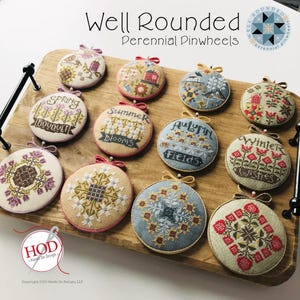 May include: A wooden tray holds twelve round, embroidered pinwheels with seasonal themes. Each pinwheel features detailed cross-stitch designs and the words "Spring," "Summer," "Autumn," and "Winter." The text "Well Rounded Perennial Pinwheels" is at the top.