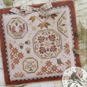 May include: A cross-stitch sampler featuring autumn-themed motifs. The design includes octagonal frames with squirrels, leaves, and floral patterns in shades of brown, green, and beige. A decorative bow and a small circular emblem with a bird are also present.