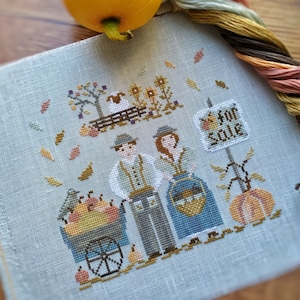 Può includere: Ricamo a punto croce con una scena di raccolta autunnale. Una coppia è in piedi vicino a un carrello pieno di zucche. Un cartello dice "in vendita". Il disegno include una pecora, girasoli e foglie che cadono. Filo da ricamo e una zucca sullo sfondo.
