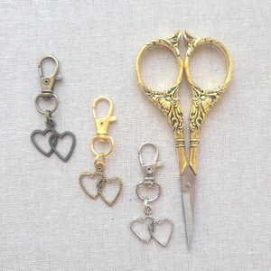 May include: An assortment of craft supplies including three heart-shaped keychains in bronze, gold, and silver tones, and a pair of ornate gold scissors. The scissors have a floral design on the handles.