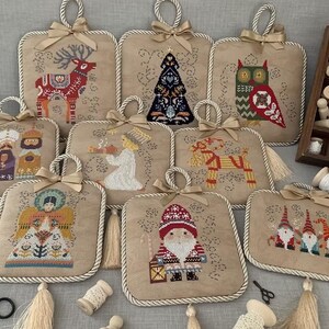 May include: A collection of embroidered Christmas-themed wall hangings. Each square piece features a unique design, including a reindeer, Christmas tree, owl, and Santa Claus. They have a beige background, cream rope trim, and a beige bow at the top.