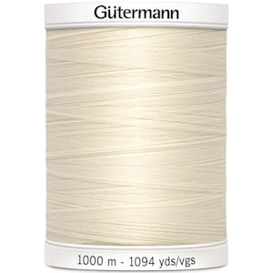 May include: A spool of Gütermann cream-colored sewing thread. The spool is cylindrical with white labels at the top and bottom. The text "Gütermann" is printed on the top label. The bottom label reads "1000 m - 1094 yds/vgs".