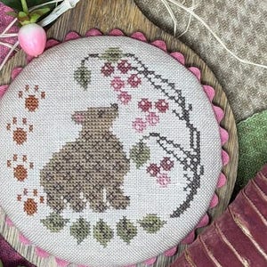 May include: A cross-stitch embroidery featuring a brown bear with a berry branch and paw prints. The embroidery is set in a round frame with a pink scalloped edge. The background includes pink and green fabric.
