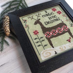 May include: Framed cross-stitch art with a Christmas theme. The artwork features a brown reindeer with a decorative pattern, surrounded by floral accents and the text "a magic little Christmas." The frame is dark brown.