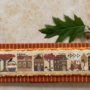 May include: A cross-stitch sampler featuring a row of autumn-themed houses, trees, and a crow, set against a beige background. The sampler is framed by a textured orange fabric. An oak leaf and acorn are placed above the sampler.