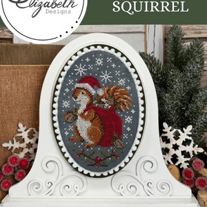 May include: A white, decorative clock with an oval cross-stitch design featuring a squirrel wearing a Santa hat and carrying a sack. The cross-stitch is framed by a green border and set against a gray background with snowflakes. The clock has a scrolled base.