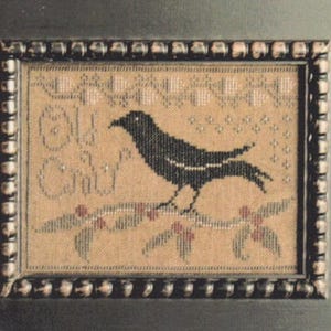 May include: A framed cross-stitch embroidery on tan fabric featuring a black bird perched on a branch with red berries and green leaves. The text "Old Crow" is stitched in black on the left side, surrounded by a decorative border in a dark frame.