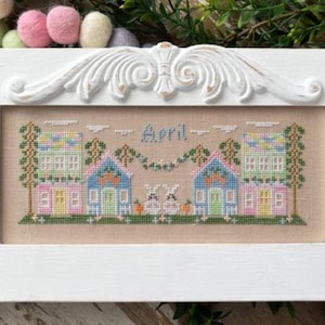 May include: A framed cross-stitch piece featuring two colorful houses with the word "April" above. The houses are flanked by trees and connected by a garland. Two bunnies and carrots are in the foreground. The frame is white.