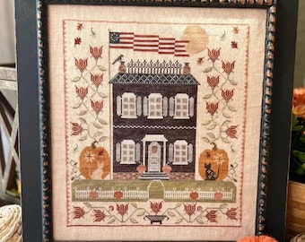 May include: Framed cross-stitch featuring a brown house with an American flag, pumpkins, and a full moon. The house has white windows and a white picket fence. The frame is black.