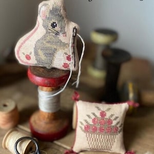 May include: A handmade, cross-stitched mouse-shaped pincushion with gray and beige details, resting on a wooden spool. A small, square pincushion with a floral basket design is also visible. The scene evokes a vintage, handcrafted aesthetic.