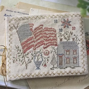 May include: A cross-stitch design depicting an American flag, a house, and floral motifs. The flag has red and white stripes with a blue star field. The house is grey with a red chimney. The design is framed with a gold border.