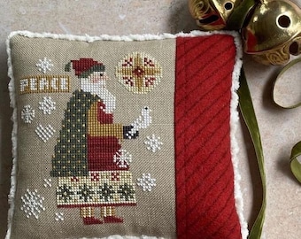 May include: A decorative Christmas pillow with a cross-stitched Santa Claus design holding a dove. The pillow features a tan background with snowflakes and the word "PEACE". A red, textured panel and white trim complete the design. Gold bells hang from a green ribbon.