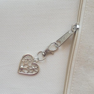 May include: A silver-colored zipper pull with a heart-shaped charm. The charm features a floral design within the heart's outline. The zipper pull is attached to a cream-colored fabric.