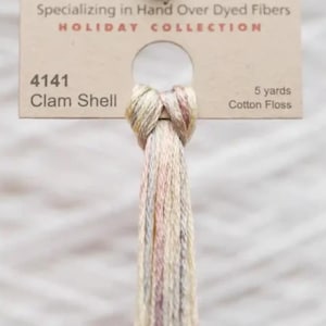 May include: A skein of cotton floss in the "Clam Shell" color, part of the Holiday Collection. The label reads "Specializing in Hand Over Dyed Fibers." The floss is a blend of pale pink, beige, and gray threads. The label also indicates 5 yards of floss.
