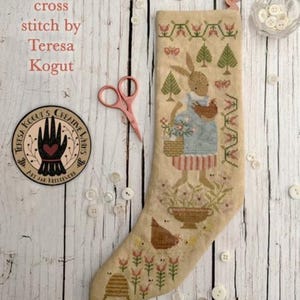 May include: A cross-stitch stocking featuring a rabbit holding a basket of flowers and a chicken. The design includes floral and geometric patterns in shades of beige, green, and red. The text "cross stitch by Teresa Kogut" is visible.