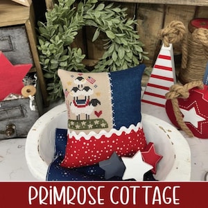 May include: A decorative pillow with a sheep design, featuring red, white, and blue colors. The pillow is in a white bowl with star-shaped ornaments. The text "PRIMROSE COTTAGE" is at the bottom of the image.