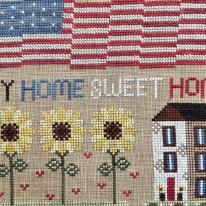 May include: Cross-stitch design featuring an American flag, the phrase "MY HOME SWEET HOME," sunflowers, and a house. The flag has red and white stripes and blue stars. The sunflowers are yellow with brown centers. The house is white with a brown roof.