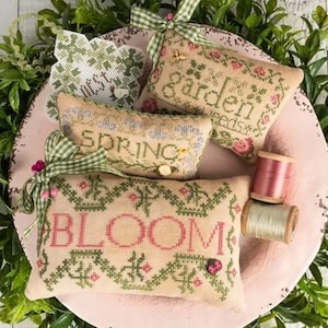 May include: A collection of embroidered decorative pillows with floral and botanical designs. The pillows feature the words "Bloom", "Spring", and "Garden Seeds" in cross-stitch. Green and pink accents, along with spools of thread, complete the spring theme.