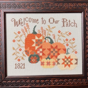 May include: A framed cross-stitch embroidery featuring the text "Welcome to Our Patch" and "1821" above and below a cluster of pumpkins with geometric star patterns in shades of orange, yellow, and green, surrounded by small sprigs and berries.