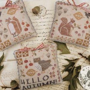 May include: Three decorative, cross-stitched pillows with autumn-themed designs. One pillow features a fox, another a squirrel, and the third says "Hello Autumn!" with a bird. Each pillow is tied with a red and white ribbon.