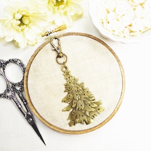 May include: A gold-toned metal Christmas tree charm with a loop for attaching to a keychain or bag. The charm is displayed on a beige fabric background.