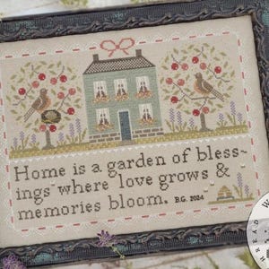 May include: Cross-stitch framed artwork featuring a house, birds, and cherry trees. The text reads "Home is a garden of blessings where love grows & memories bloom." The frame is dark with a distressed finish.