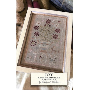 May include: A framed cross-stitch pattern with a floral design in shades of red, brown, and green. The pattern is titled "JOY" and is a 2023 Nashville Exclusive by Shakespeare's Peddler.
