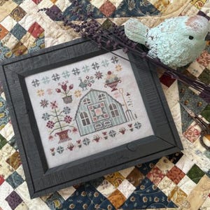 May include: A framed cross-stitch featuring a greenhouse design with floral and garden elements in shades of blue, red, and green. The frame is dark gray, and the artwork is displayed on a patterned quilt. A decorative bird and lavender sprigs are nearby.