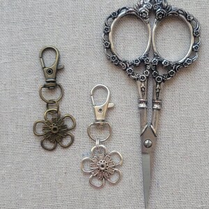 May include: An assortment of craft supplies, including a pair of ornate silver scissors and two flower-shaped keychains. The scissors have a detailed floral design on the handles. One keychain is silver, the other is bronze.