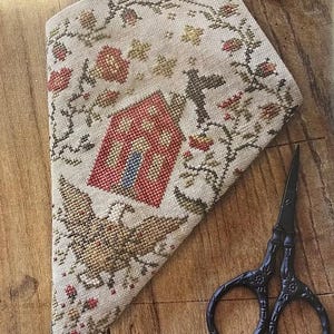 May include: A beige, cross-stitched needlework piece featuring a house and eagle design, framed by floral patterns. A pair of black scissors sits on the wooden surface beside the needlework. The needlework has a red tassel.