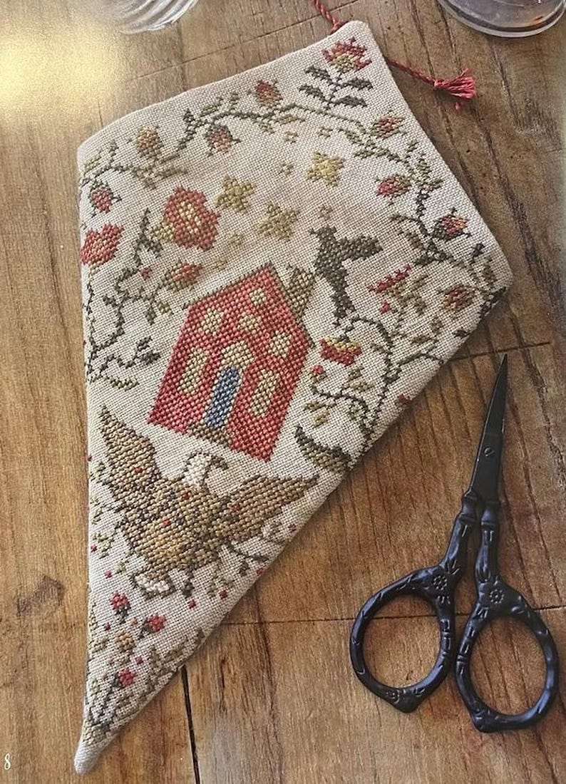 May include: A beige, cross-stitched needlework piece featuring a house and eagle design, framed by floral patterns. A pair of black scissors sits on the wooden surface beside the needlework. The needlework has a red tassel.