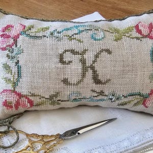 May include: A rectangular, off-white, embroidered pillow with a floral design and the letter "K". The embroidery features pink roses, green leaves, and blue accents. Gold scissors and a decorative cord are in the foreground.