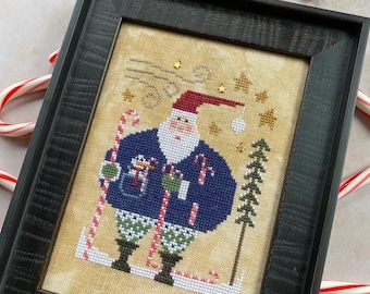 May include: Framed cross-stitch art featuring Santa Claus skiing, holding candy canes, and wearing a blue sweater and red hat. The artwork includes a Christmas tree and star accents. The frame is black, and candy canes surround the artwork.