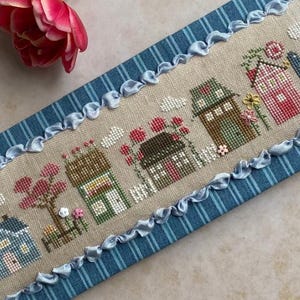 May include: A cross-stitch sampler featuring a row of colorful houses with flowers and trees. The houses are in shades of blue, pink, green, and yellow. The sampler is framed with a blue striped fabric and a ruffled ribbon.