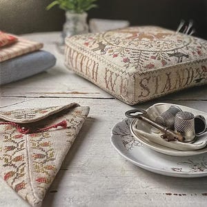 May include: A selection of vintage items on a white wooden surface. Features a stitched cushion with the words "WE STAND", a small pouch with a floral pattern, a key, and thimbles on a decorative plate. The cushion has a circular deer design.