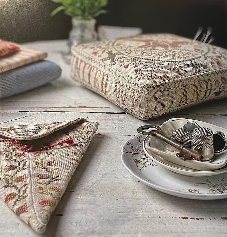 May include: A selection of vintage items on a white wooden surface. Features a stitched cushion with the words "WE STAND", a small pouch with a floral pattern, a key, and thimbles on a decorative plate. The cushion has a circular deer design.