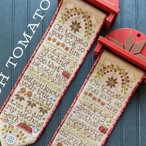 May include: Two cross-stitch banners with red wooden holders. The banners feature the text "Oh tomato you look so fine growing on that blessed vine." The design includes tomatoes, leaves, and a star. The banners are on a blue background.