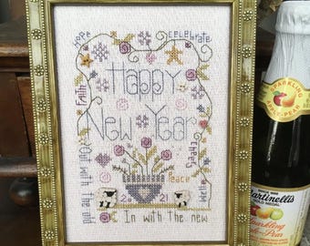 May include: A framed cross-stitch art piece with the words "Happy New Year" and other text, surrounded by floral and snowflake designs. A bottle of sparkling apple-pear juice is next to the frame.