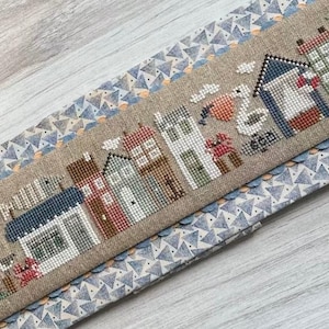 May include: A cross-stitch design featuring a seaside town scene with houses, a lighthouse, and seagulls. The design is stitched on a tan fabric and framed by a blue and white patterned border. The word "sea" is stitched into the design.