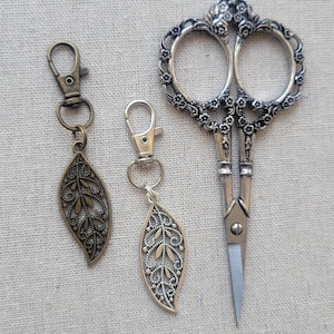 May include: Two metal leaf-shaped charms attached to lobster claw clasps and a pair of ornate silver-toned scissors with floral detailing on the handles. The leaf charms feature intricate cut-out patterns, with one in a bronze tone and one in silver.
