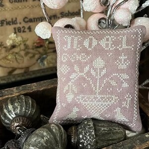 May include: A small, rectangular, lavender-colored decorative pillow with white cross-stitch embroidery. The embroidery features the word "NOEL", floral designs, and geometric patterns. The pillow is displayed in a wooden box with vintage ornaments.