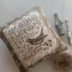 May include: Square decorative pillow with a stitched bird design and the word "Cuckoo" and the year 2023. The pillow has a light beige center with a brown bird and alphabet design. The border is olive green with a white paisley pattern.