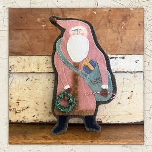 May include: A felt Santa Claus figure in shades of pink, white, and green. The figure has a long coat, a white beard, and a bag labeled "toys." He carries a green wreath and a candy cane. The figure is set against a rustic wooden background.