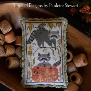 May include: A cross-stitch pillow with a raccoon family design. The pillow features a green border and a cream-colored background with corn stalks and a pumpkin. The text "Original Designs by Paulette Stewart" is at the top.