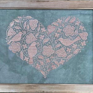 May include: Framed cross-stitch artwork featuring a heart-shaped design filled with pink birds, flowers, and foliage on a teal background. The artwork is enclosed in a light brown wooden frame, creating a rustic aesthetic.